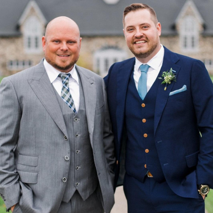 Two men smile towards the camera. One is wearing a grey suit and vest, and the other is wearing a dark blue suit and vest.