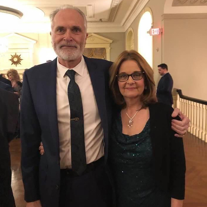 A man and woman stand arm in arm, smiling towards the camera. He is wearing a black suit and tie.